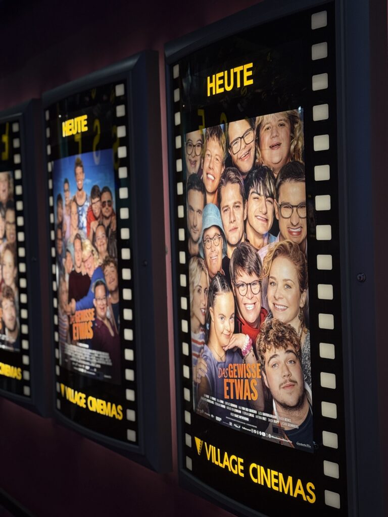 Auf zwei Filmplakaten in deutscher Sprache ist eine Gruppe lächelnder Menschen zu sehen. Die Plakate sind in beleuchteten Rahmen in einem Kino angebracht, wobei oben und unten die Schriftzüge „HEUTE“ und „VILLAGE CINEMAS“ zu sehen sind.