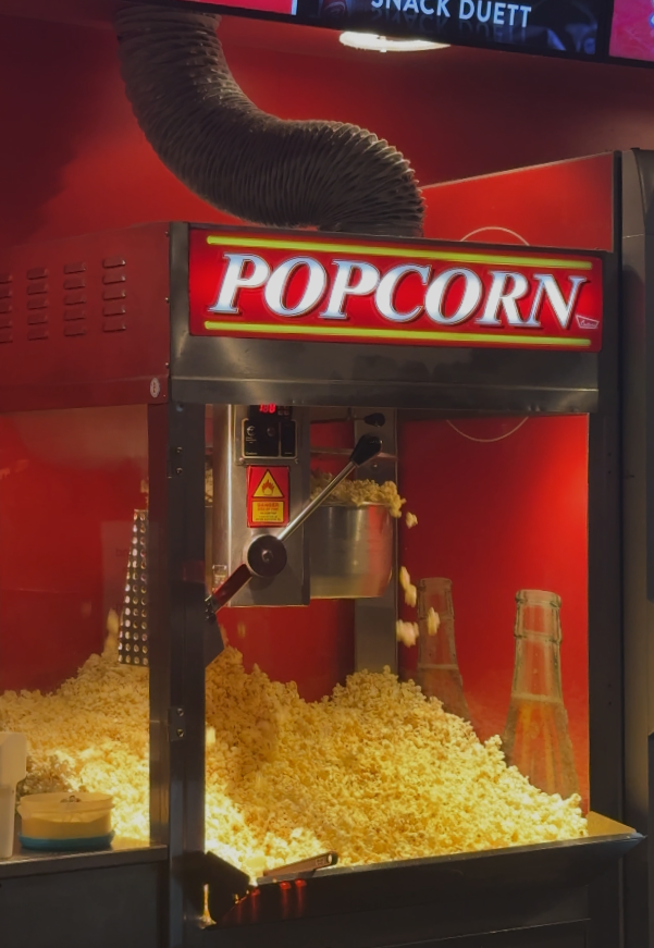 Eine Popcornmaschine quillt über vor frisch zubereitetem Popcorn, das die Schale füllt und auf die darunterliegende Fläche tropft. Hinter dem Popcornberg stehen zwei durchsichtige Flaschen. Die Maschine und der Hintergrund sind leuchtend rot.