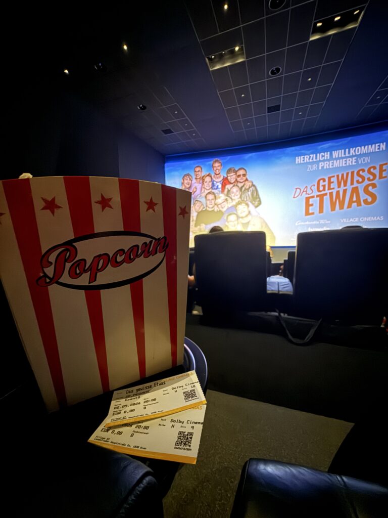 Eine gestreifte Popcorn-Schachtel und Kinokarten in einem Kino, in dem Menschen sitzen und auf der Leinwand die Aufschriften „Das gewisse Etwas“ und „Herzlich Willkommen zur Premiere“ auf Deutsch zu sehen sind.