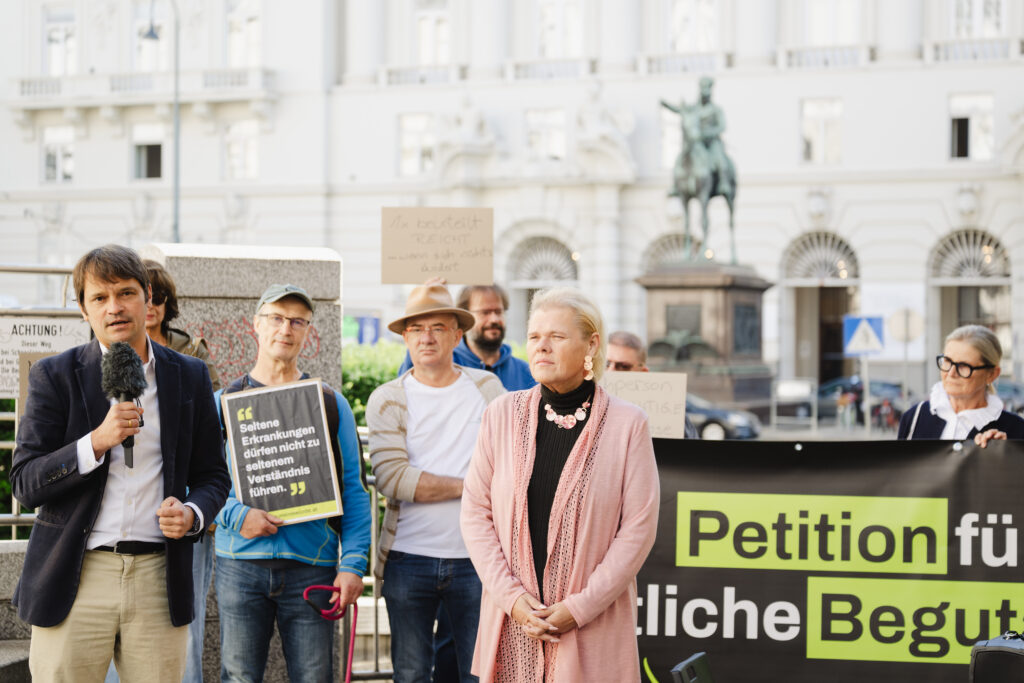 Eine Gruppe von Menschen steht im Freien und hält Schilder sowie ein Transparent mit der Aufschrift „Petition für…“ hoch, während ein Mann in ein Mikrofon spricht. Im Vordergrund steht eine Frau in einem rosa Pullover. Im Hintergrund sind Gebäude und eine Statue zu sehen.