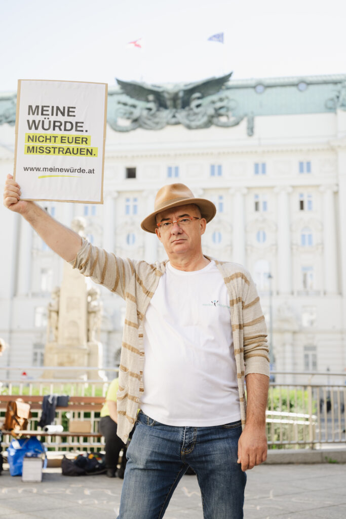 Ein Mann mit Hut und Strickjacke hält auf einem Stadtplatz ein Schild mit der Aufschrift „MEINE WÜRDE. NICHT EUER MISSTRAUEN.“ in der Hand, im Hintergrund ist ein historisches Gebäude zu sehen.
