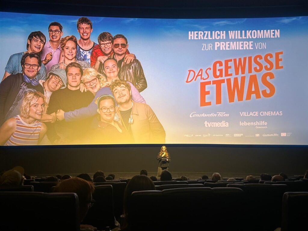 Eine Frau steht auf der Bühne eines Kinos und spricht zu den Zuschauern. Hinter ihr ist auf einer großen Leinwand ein Gruppenfoto und ein Text zu sehen, mit denen für den Film „Das Gewisse Etwas“ anlässlich seiner Premiere geworben wird.