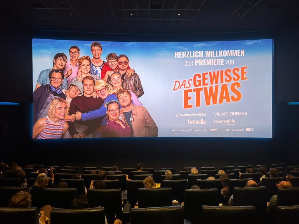 Die Zuschauer sitzen in einem Kino und blicken auf eine Leinwand, auf der eine Begrüßungsbotschaft zur Premiere von „Das Gewisse Etwas“ zu sehen ist, zusammen mit einem großen Gruppenfoto der lächelnden Darsteller vor blauem Hintergrund.