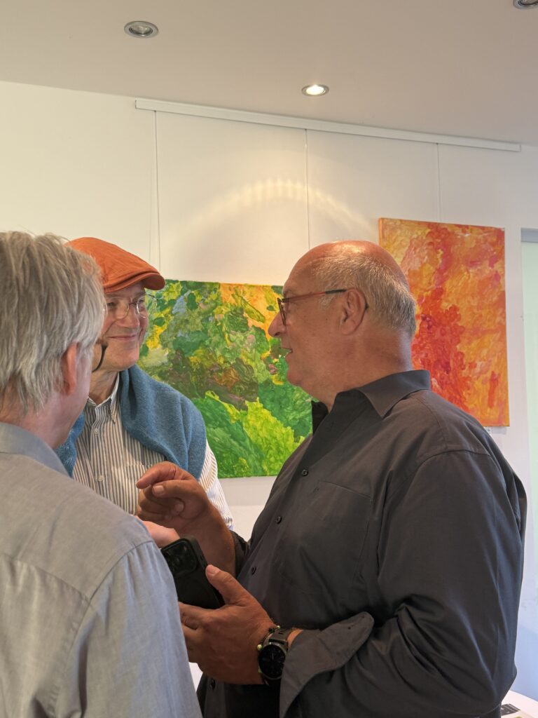 Drei Männer unterhalten sich in einer Kunstgalerie, hinter ihnen hängen farbenfrohe abstrakte Gemälde an der Wand. Ein Mann trägt eine orangefarbene Mütze, ein anderer hält ein Handy in der Hand, und alle scheinen in ein freundliches Gespräch vertieft zu sein.