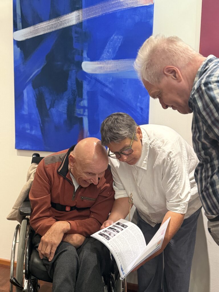 Drei ältere Menschen, von denen einer im Rollstuhl sitzt, schauen sich gemeinsam eine Zeitschrift oder eine Broschüre an. Sie wirken gebannt und interessiert. An der Wand hinter ihnen hängt ein großes blaues abstraktes Gemälde.