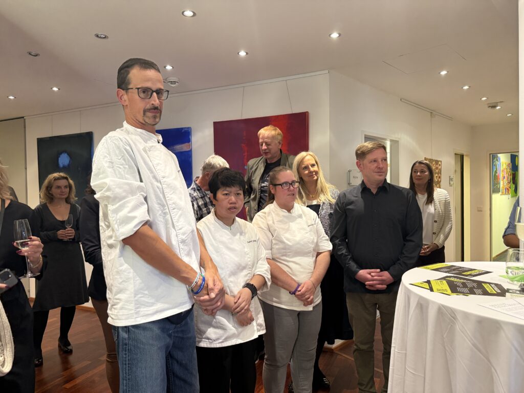 Eine Gruppe von Menschen, darunter Köche in weißen Uniformen, steht gemeinsam bei einer Veranstaltung in einem Saal, im Hintergrund sind farbenfrohe Kunstwerke an den Wänden zu sehen. Einige Personen hören aufmerksam an einem runden Tisch zu.