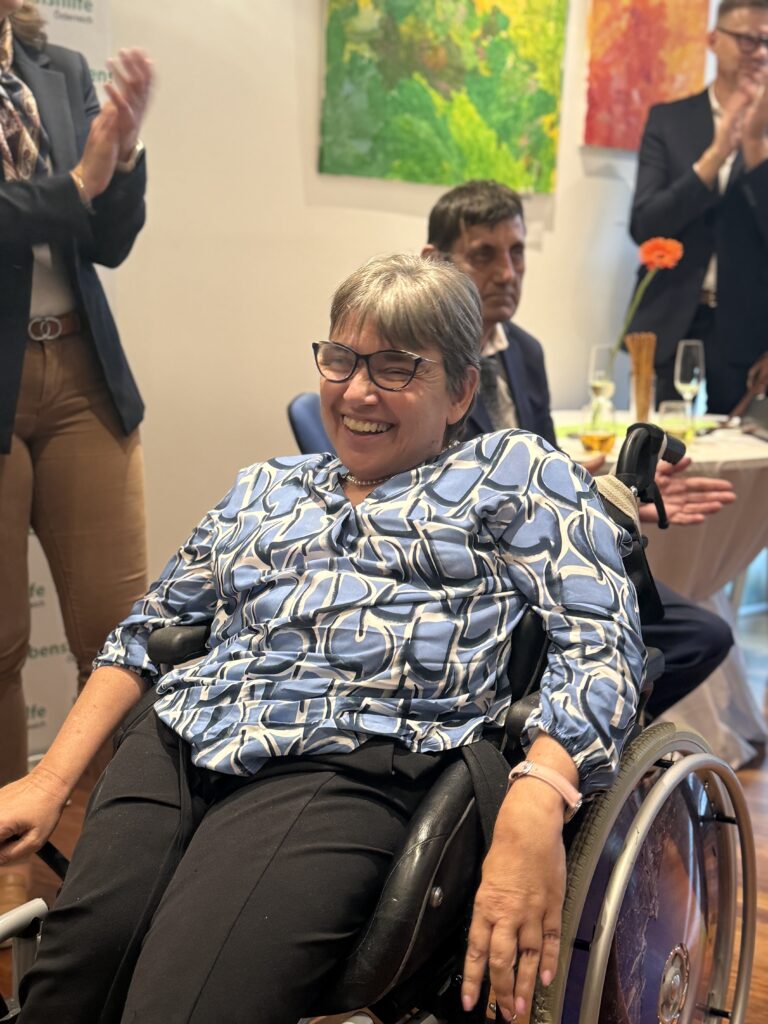 Eine lächelnde Frau im Rollstuhl, die eine Brille und ein gemustertes blaues Hemd trägt, befindet sich bei einer Veranstaltung in einem Saal, während hinter ihr Menschen klatschen und bunte Kunstwerke an der Wand hängen.