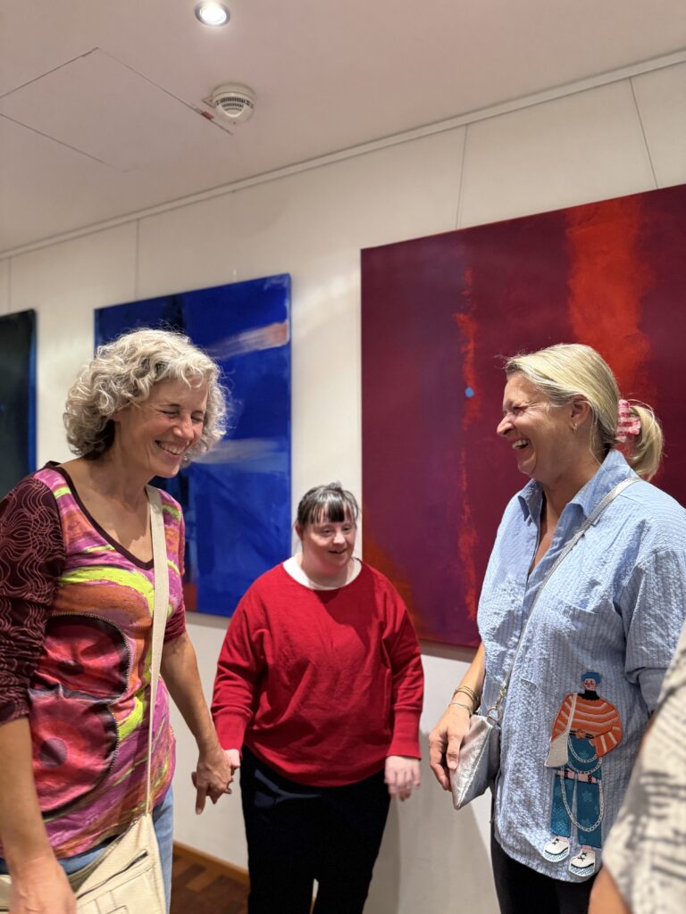 Drei Frauen stehen in einer Kunstgalerie, lächeln und unterhalten sich. Zwei von ihnen stehen im Vordergrund und halten sich an den Händen, eine steht zwischen ihnen. An der weißen Wand hinter ihnen hängen farbenfrohe abstrakte Gemälde.