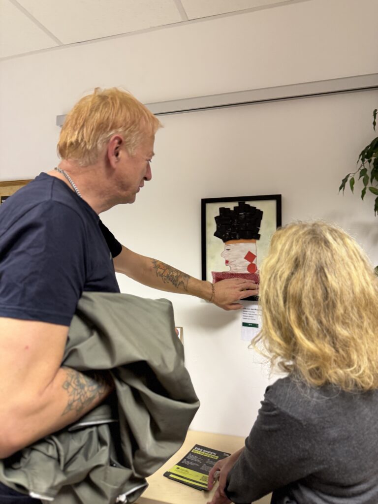 Ein Mann und eine Frau betrachten in einem Innenraum ein gerahmtes Wandbild und unterhalten sich darüber. Der Mann zeigt auf das Kunstwerk und hält eine Jacke in der Hand, während die Frau daneben steht. Auf dem Tisch liegen Informationsbroschüren.