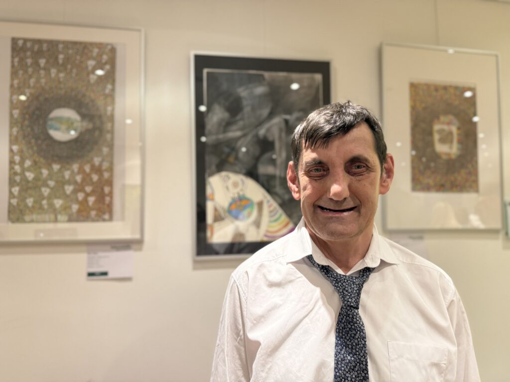 Ein Mann in einem weißen Hemd und einer gemusterten Krawatte steht lächelnd vor drei gerahmten Kunstwerken, die an einer Galeriewand ausgestellt sind.