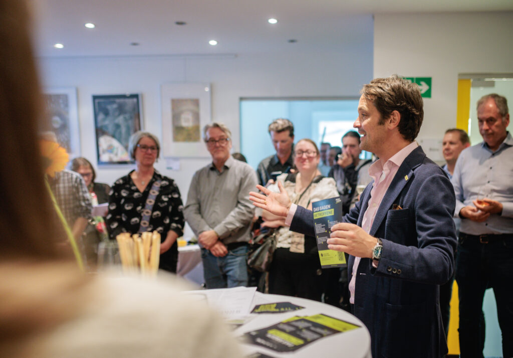 Ein Mann in einem blauen Blazer und einem hellrosa Hemd hält eine Broschüre mit dem Titel DAS SAGEN BETROFFENE in der Hand, während er zu einer bunt gemischten Gruppe aufmerksamer Zuhörer spricht, die sich in einem hell beleuchteten Raum versammelt haben.
