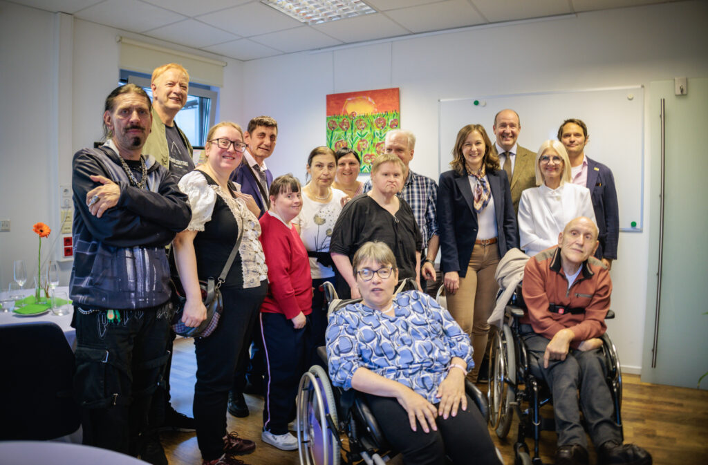 Eine Gruppe von Menschen, darunter auch Personen im Rollstuhl, posiert gemeinsam und lächelt in einem hell beleuchteten Raum mit Tischen und Kunstwerken an den Wänden.