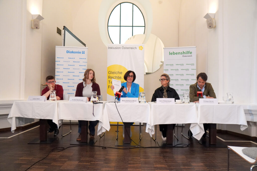 Fünf Personen sitzen während einer Podiumsdiskussion an einem langen Tisch, im Hintergrund sind Werbebanner zu sehen.