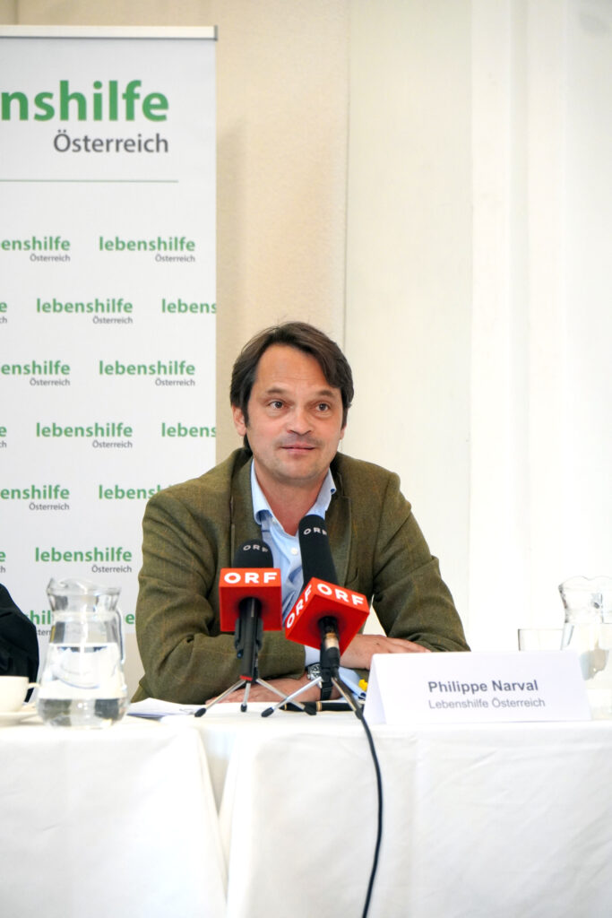 Philippe Narval von der Lebenshilfe Österreich spricht auf einer Pressekonferenz, vor ihm stehen zwei Mikrofone.