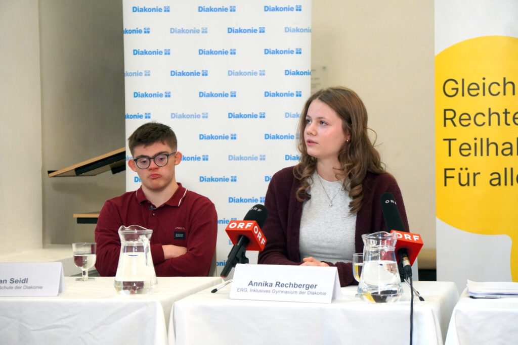 Ein junger Mann und eine junge Frau sitzen während einer Pressekonferenz vor einem Diakonie-Banner an einem Tisch mit Mikrofonen.