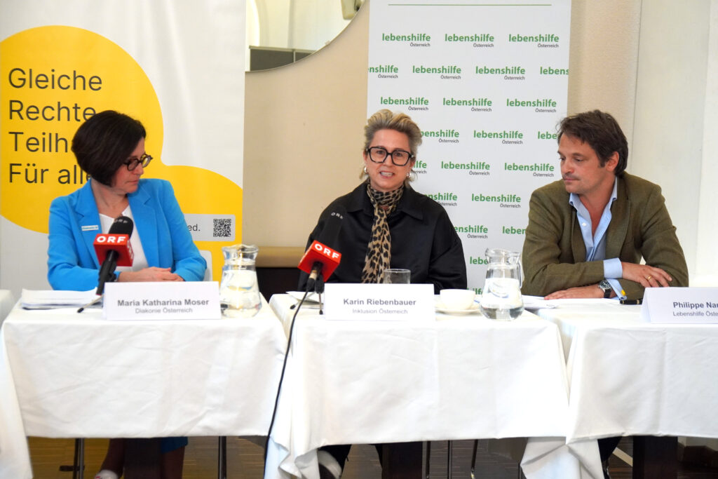 Drei Personen sitzen während einer Pressekonferenz an einem Tisch, vor ihnen stehen Mikrofone und Namensschilder mit den Namen Maria Katharina Moser, Karin Riebenbauer und Philippe Narval.
