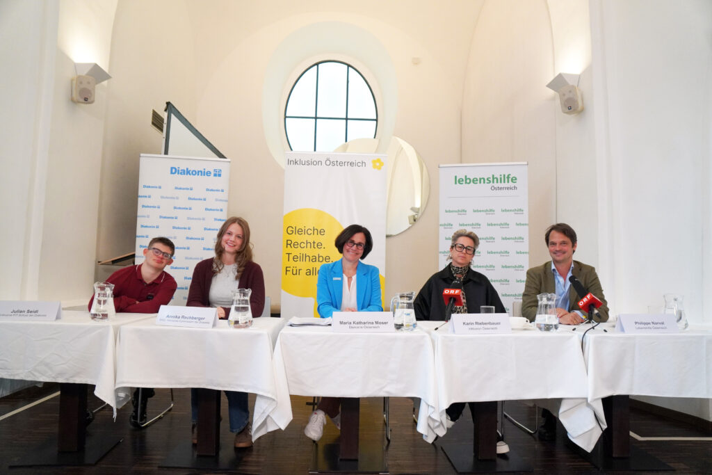 Fünf Personen sitzen bei einer Pressekonferenz hinter einem langen Tisch, im Hintergrund sind Banner der Diakonie und der Lebenshilfe zu sehen.