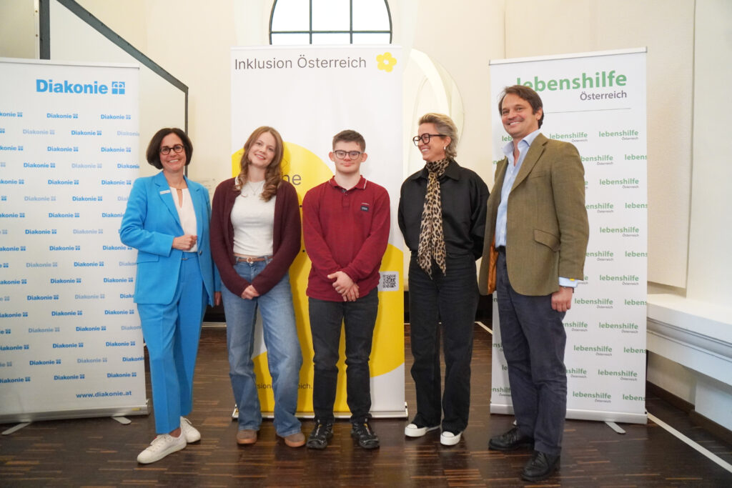 Fünf Personen stehen in einer Reihe und posieren für ein Foto vor drei hochformatigen Bannern der Diakonie, von Inklusion Österreich und der Lebenshilfe.