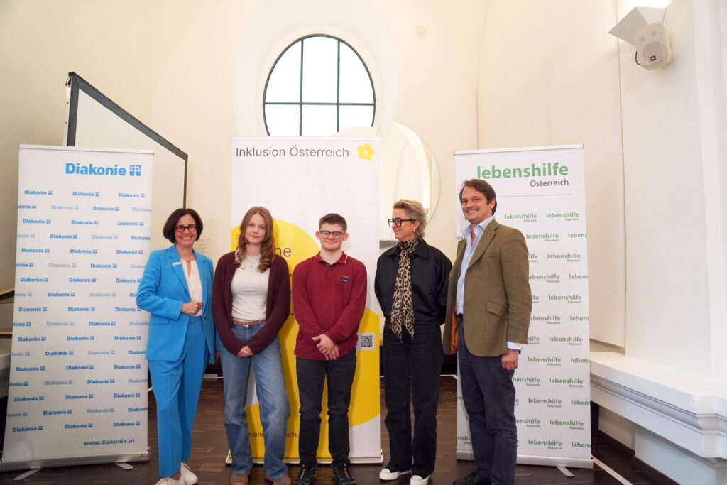 Fünf Personen stehen in einer Reihe vor zwei Werbebannern der Diakonie und der Lebenshilfe Österreich.