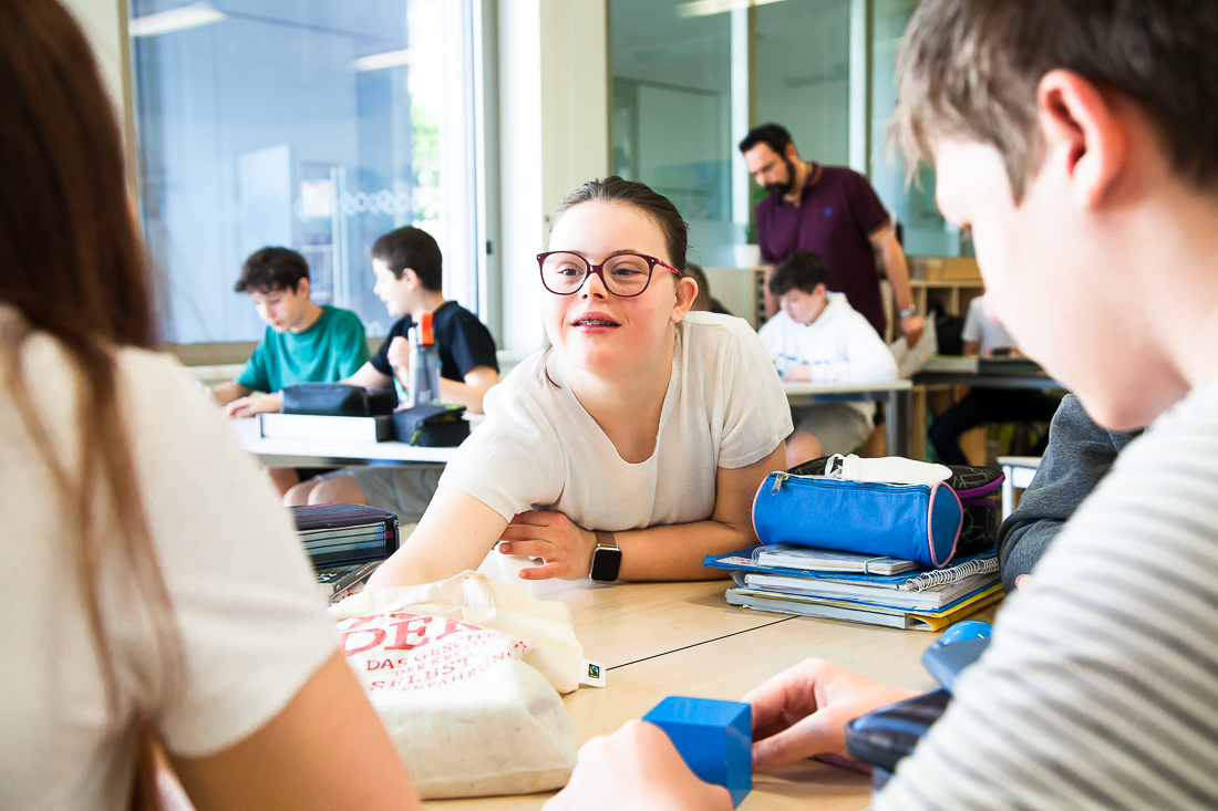 Eine junge Frau mit Brille beugt sich über einen Tisch, um sich mit ihren Klassenkameraden in einem hellen, modernen Klassenzimmer zu unterhalten.