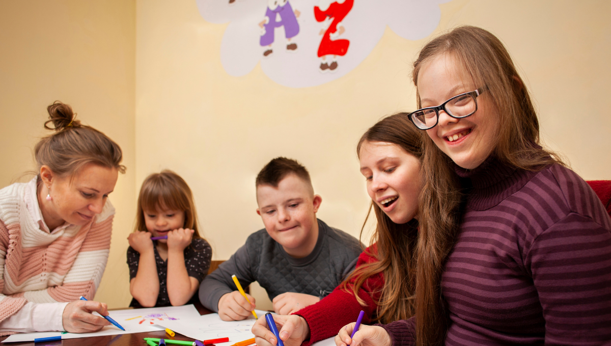 Eine Lehrkraft und vier Kinder sitzen gemeinsam an einem Tisch und malen – ein Bild, das den Auftrag von Lebenshilfe Österreich widerspiegelt, Inklusion und Teilhabe zu fördern.